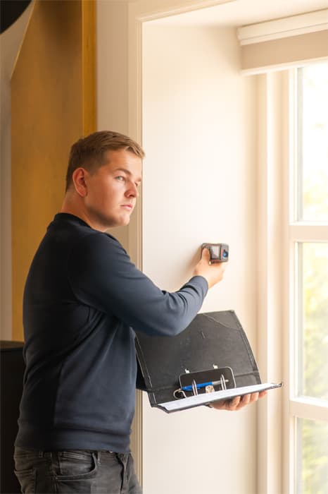 Jan-thijs meet met een apparaat de muur bij een raam op en houdt een zwarte ordner vast.